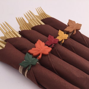 Puede incluir: Cinco servilletas marrones con tenedores dorados envueltos alrededor. Cada servilleta tiene una hoja de otoño de color diferente atada alrededor. Las hojas son verdes, naranjas, rojas, amarillas y marrones.
