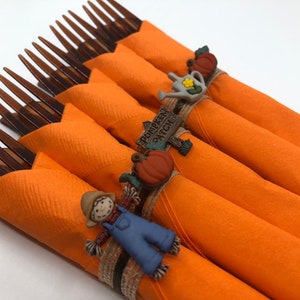 Puede incluir: Servilletas naranjas con anillos de servilleta de temática otoñal. Los anillos presentan un espantapájaros, una calabaza y un letrero que dice "Pumpkin Patch".