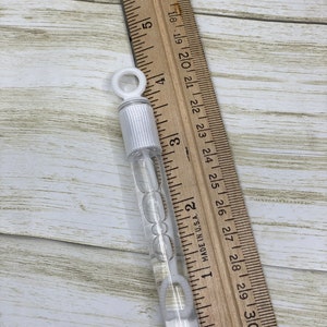 Puede incluir: Una varita de burbujas de pl&aacute;stico transparente con un anillo blanco en la parte superior. La varita mide aproximadamente 15 cm de largo y est&aacute; llena de soluci&oacute;n de burbujas transparente. La varita est&aacute; sobre una regla de madera.
