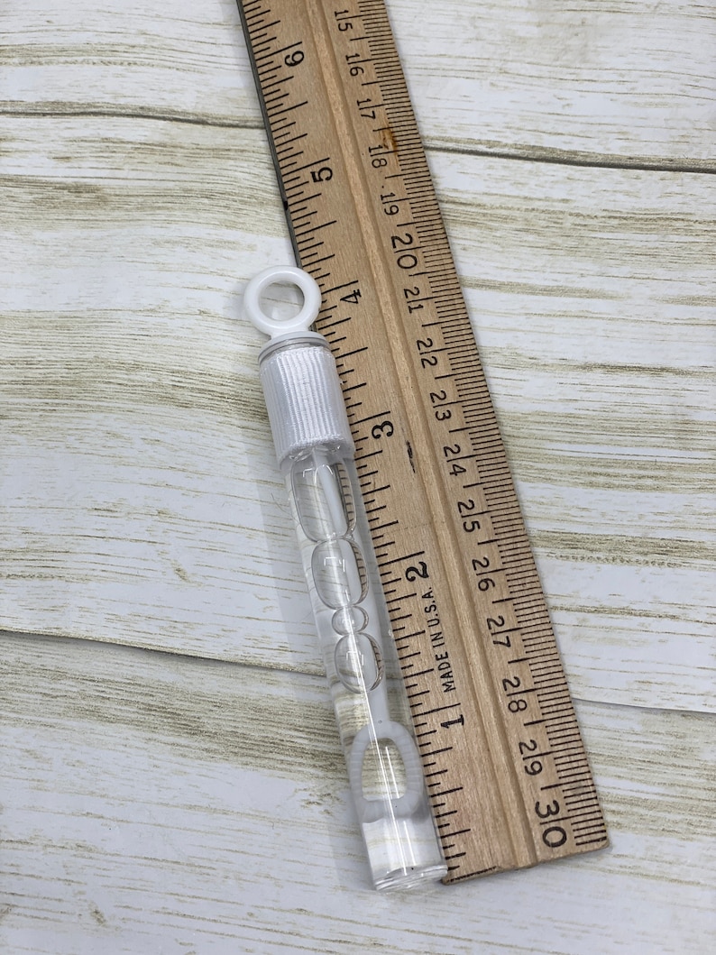 Puede incluir: Una varita de burbujas de pl&aacute;stico transparente con un anillo blanco en la parte superior. La varita mide aproximadamente 15 cm de largo y est&aacute; llena de soluci&oacute;n de burbujas transparente. La varita est&aacute; sobre una regla de madera.