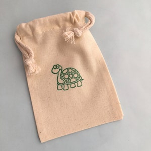 May include: A beige drawstring bag with a green embroidered turtle design. The bag is made of a natural-colored fabric and features a simple, cartoon-style turtle. The drawstring is a matching beige color.