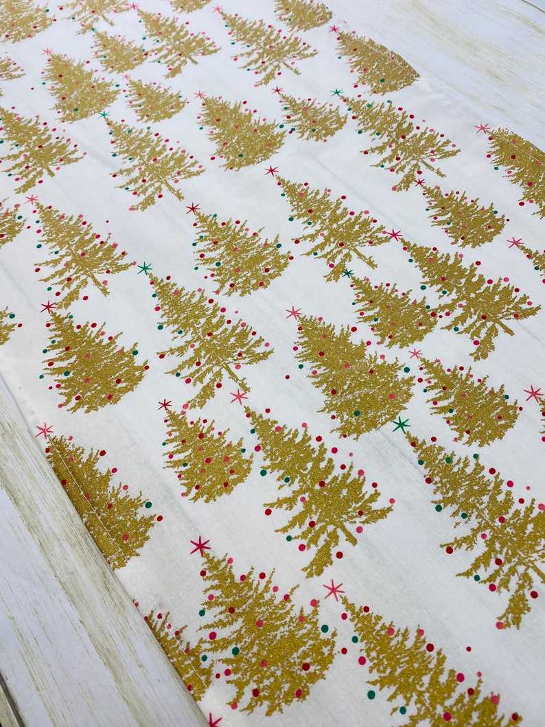 May include: White table runner with a gold glitter Christmas tree pattern. The trees are decorated with red and green ornaments.