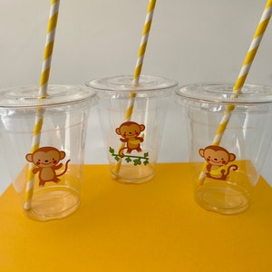 以下が含まれることがあります： 黄色と白のストライプのストローが付いた透明なプラスチックカップが3つ。各カップにはバナナを持った猿のステッカーが貼られています。カップは黄色の表面の上に置かれています。
