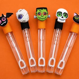 May include: Five clear plastic bubble wands featuring Halloween-themed toppers. The toppers include a black bat, a white mummy, a green Frankenstein monster, a white skull, and a black cat. Each wand has a colored cap and a white bubble wand inside.