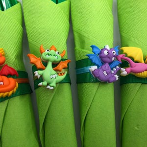May include: Four green napkins with dragon-shaped napkin rings. The dragons are orange, green, purple, and pink.