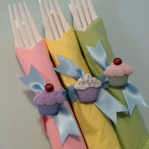 May include: Three white plastic forks wrapped in colorful napkins with blue ribbon and cupcake-shaped napkin rings. The napkins are pink, yellow, and green.