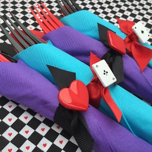 May include: Four forks wrapped in purple and turquoise napkins with red and black ribbon and white playing card accents. The napkins are on a black and white checkered surface.
