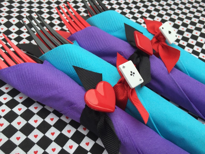 May include: Four forks wrapped in purple and turquoise napkins with red and black ribbon and white playing card accents. The napkins are on a black and white checkered surface.