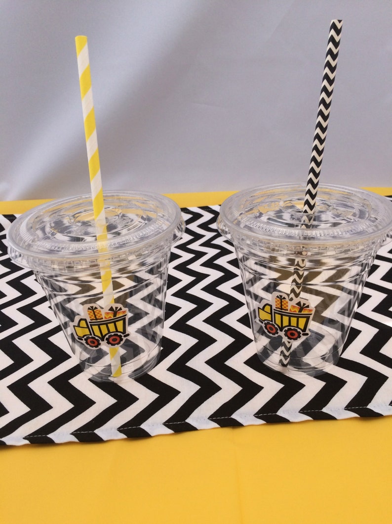 May include: Two clear plastic cups with black and white chevron patterned lids. Each cup has a yellow construction truck sticker and a striped straw. The cups are on a black and white chevron patterned tablecloth.