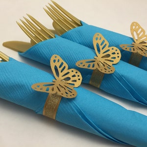 May include: Three blue napkins with gold butterfly napkin rings. The napkins are rolled up and the forks are placed inside the napkins.