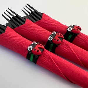 May include: Three red napkins with black forks wrapped inside. Each napkin has a ladybug napkin ring with black and green stripes and white googly eyes.