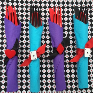 May include: Four sets of silverware wrapped in napkins, tied with red ribbon and a playing card charm. The napkins are purple, blue, and white with a black and white checkered background.