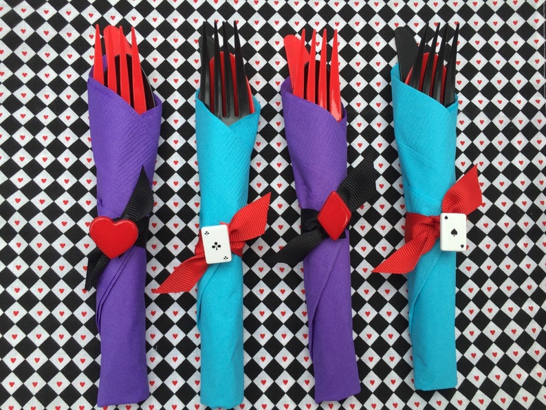 May include: Four sets of silverware wrapped in napkins, tied with red ribbon and a playing card charm. The napkins are purple, blue, and white with a black and white checkered background.