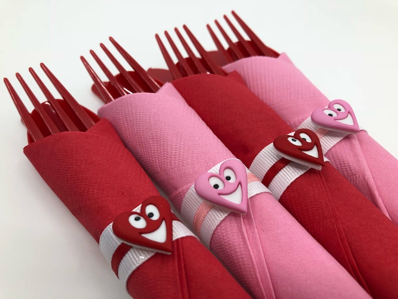 May include: Four red plastic forks wrapped in pink and red napkins with white ribbon and red heart-shaped decorations with smiley faces.