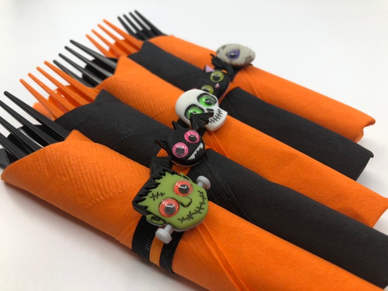 May include: Set of five Halloween-themed napkin rings. Each ring features a different spooky character, including a skull, a cat, a monster, and a ghost. The rings are made of black and orange fabric and are decorated with googly eyes.