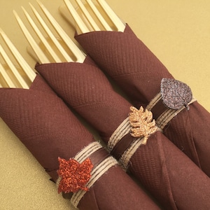 Puede incluir: Tres servilletas marrones están enrolladas y atadas con cuerda y una hoja brillante. Las hojas son doradas, marrones y rojas.