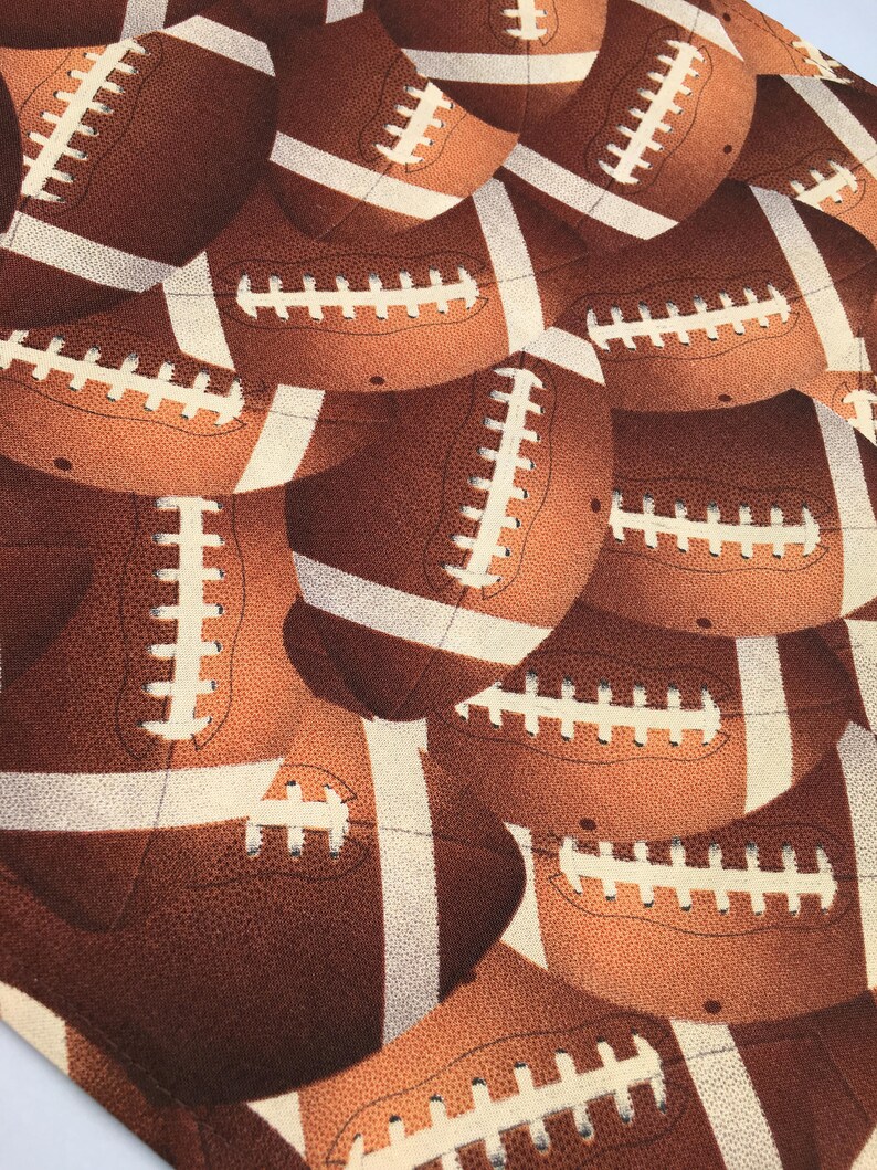 May include: A close-up of a fabric with a repeating pattern of brown and white footballs. The footballs have white laces and are arranged in a staggered pattern.