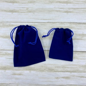 May include: Two blue velvet drawstring pouches, one larger than the other, on a white wooden surface.