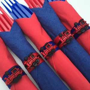 May include: Red and blue napkins with train-shaped napkin rings. The rings are black, red, and blue.