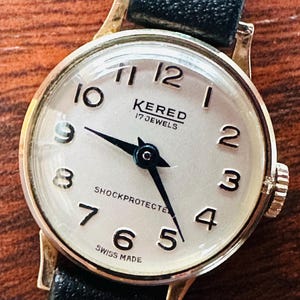 May include: A vintage Kered wristwatch featuring a gold-coloured case and a black leather strap. The watch face is white with black numerals and hands. The text "KERED 17 JEWELS" and "SHOCKPROTECTED" are visible. The watch is marked "SWISS MADE".