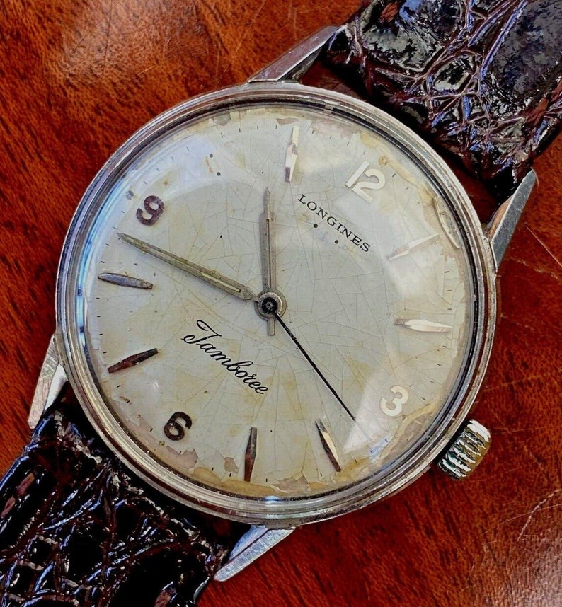 May include: A silver Longines watch with a white face and black hands. The watch has a brown leather band and the words "Longines" and "Jamboree" printed on the face.