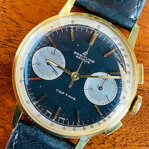 May include: A gold-tone Breitling Geneve Top Time chronograph wristwatch with a black face and white subdials. The watch has a black leather band.