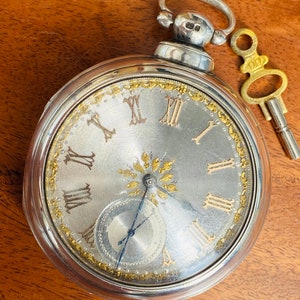 Könnte beinhalten: Eine silberne Taschenuhr mit weißem Zifferblatt und goldenen römischen Ziffern für die Stunden. Die Uhr hat ein dekoratives goldenes Design am Rand des Zifferblatts und ein kleines, rundes, silbernes Hilfszifferblatt. Die Uhr ist an einer silbernen Kette mit einem Schlüssel befestigt.