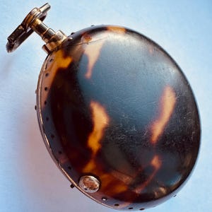 Könnte beinhalten: Eine antike Taschenuhr mit Schildpattmuster auf dem Gehäuse. Die Uhr hat einen goldfarbenen Aufzugsstift und einen kleinen, kreisförmigen Knopf. Das ovale Gehäuse hat eine dunkelbraune und bernsteinfarbene Farbgebung.