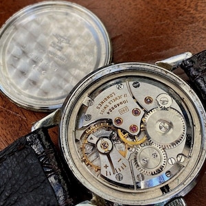 May include: Close-up of the inner workings of a silver Longines watch with a 17-jewel movement. The watch face is visible with the text "Longines 17 Jewels Swiss 10646624 280".