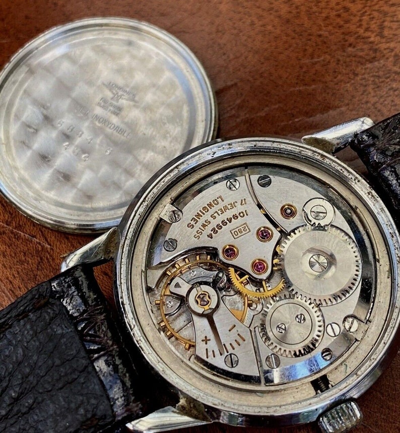 May include: Close-up of the inner workings of a silver Longines watch with a 17-jewel movement. The watch face is visible with the text "Longines 17 Jewels Swiss 10646624 280".