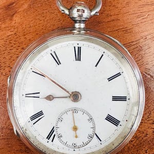 Könnte beinhalten: Eine silberne Taschenuhr mit weißem Zifferblatt, schwarzen römischen Ziffern und einem kleinen Sekundenzeiger. Die Uhr hat eine silberne Schlaufe oben und eine detaillierte silberne Lünette. Die Zeiger sind schwarz und kupferfarben.