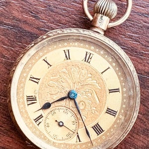 Könnte beinhalten: Eine goldfarbene Taschenuhr mit rundem Zifferblatt, schwarzen römischen Ziffern, einer zentralen Blumengravur und einem kleinen Hilfszifferblatt für Sekunden. Die Uhr hat eine Aufzugskrone oben mit einer runden Öse zur Befestigung.