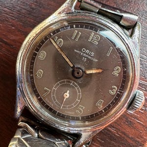 Puede incluir: Reloj de pulsera Oris impermeable vintage con caja plateada y esfera gris oscuro. La esfera presenta números arábigos, una subesfera y el texto "ORIS WATERPROOF 15". El reloj tiene una correa de metal plateado.