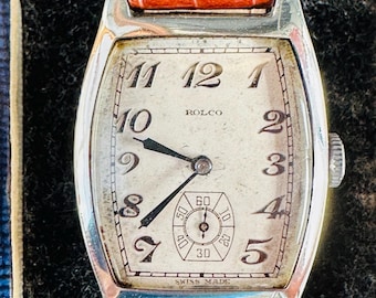 May include: A silver wristwatch with a white face and black hands. The watch has a brown leather band and says "ROLCO" and "SWISS MADE" on the face.