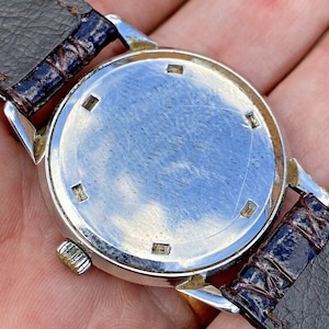 May include: A silver wristwatch with a brown leather band. The watch face is covered with a clear glass cover. The back of the watch is visible, showing a smooth, silver surface with a few small marks.