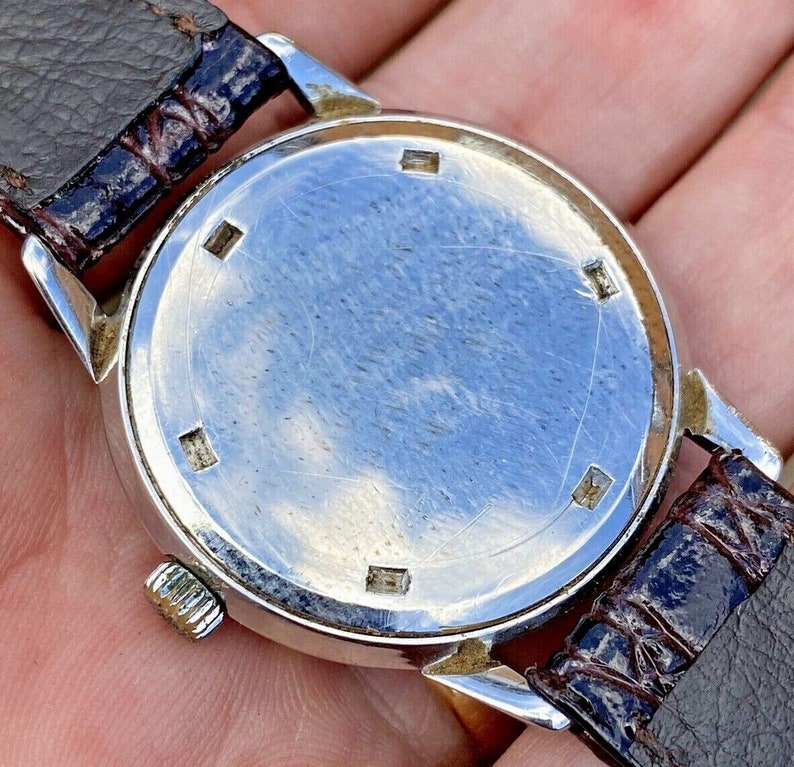 May include: A silver wristwatch with a brown leather band. The watch face is covered with a clear glass cover. The back of the watch is visible, showing a smooth, silver surface with a few small marks.