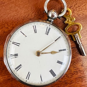 May include: A silver pocket watch with Roman numeral hour markers and a key attached. The watch face is white with black hands.