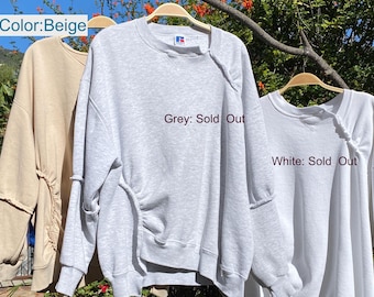 May include: Three sweatshirts in beige, gray, and white hang on wooden hangers. The gray and white sweatshirts have the text "Sold Out" printed on them. The beige sweatshirt is on the left, the gray in the middle, and the white on the right.
