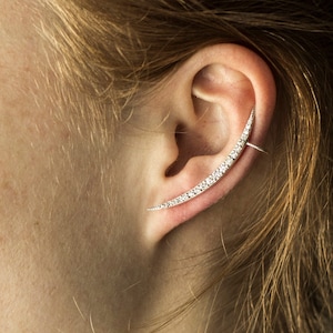 May include: A close-up of a person's ear with a silver ear cuff adorned with small, clear crystals. The ear cuff is curved and extends from the earlobe to the top of the ear.