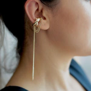 May include: Gold-toned ear cuff featuring a bird design with dangling chains. The ear cuff wraps around the ear and has a long chain that hangs down. The jewelry is worn on the ear.