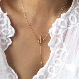 Può includere: Una delicata collana d'oro con un design a stella e un pendente a croce pendente. La collana è adornata con piccole pietre preziose scintillanti. La persona indossa una camicetta bianca ricamata.