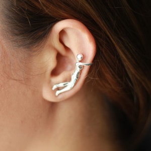 May include: Silver ear cuff featuring a stylized human figure design. The figure is positioned in a reclining pose, with one arm extended and the other bent at the elbow. The cuff is designed to wrap around the upper ear.