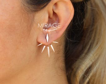 May include: A pair of gold-toned earrings with a starburst design. The earrings are made of metal and have a sharp, pointed design. The earrings are worn in the ear of a person with blonde hair.