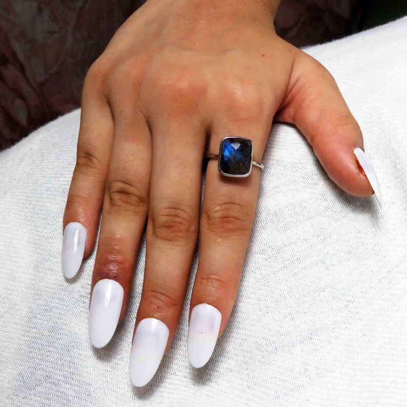May include: A silver ring with a square, dark blue gemstone is worn on a finger. The hand has long, white, oval-shaped fingernails. The ring is the focal point, showcasing the gemstone's color and shape.