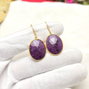 May include: A pair of gold-colored drop earrings featuring oval-shaped, faceted purple gemstones. The earrings have a gold-colored frame and a simple hook design. The gemstones have a speckled appearance.