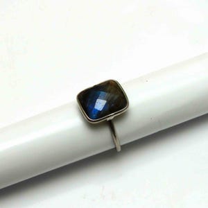 May include: A silver ring featuring a square-cut, iridescent labradorite gemstone. The stone displays shades of blue, green, and brown. The ring's band is a simple, thin silver design, showcasing the gemstone's unique color.