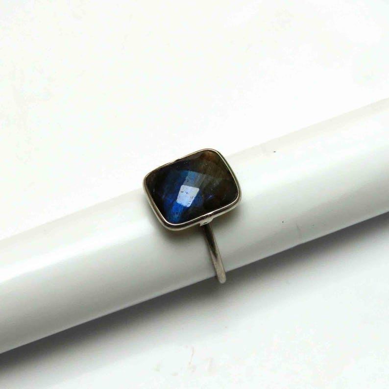 May include: A silver ring featuring a square-cut, iridescent labradorite gemstone. The stone displays shades of blue, green, and brown. The ring's band is a simple, thin silver design, showcasing the gemstone's unique color.