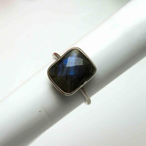 May include: A silver ring featuring a faceted, rectangular labradorite gemstone. The gemstone displays iridescent blue and green hues. The ring is set on a white background, showcasing the jewelry's design and color.