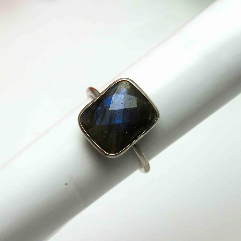 May include: A silver ring featuring a faceted, rectangular labradorite gemstone. The gemstone displays iridescent blue and green hues. The ring is set on a white background, showcasing the jewelry's design and color.
