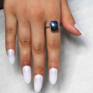 May include: A silver ring with a square, faceted, dark blue gemstone. The ring is on a hand with long, white, oval-shaped fingernails. The gemstone has a reflective quality, showing flashes of light.
